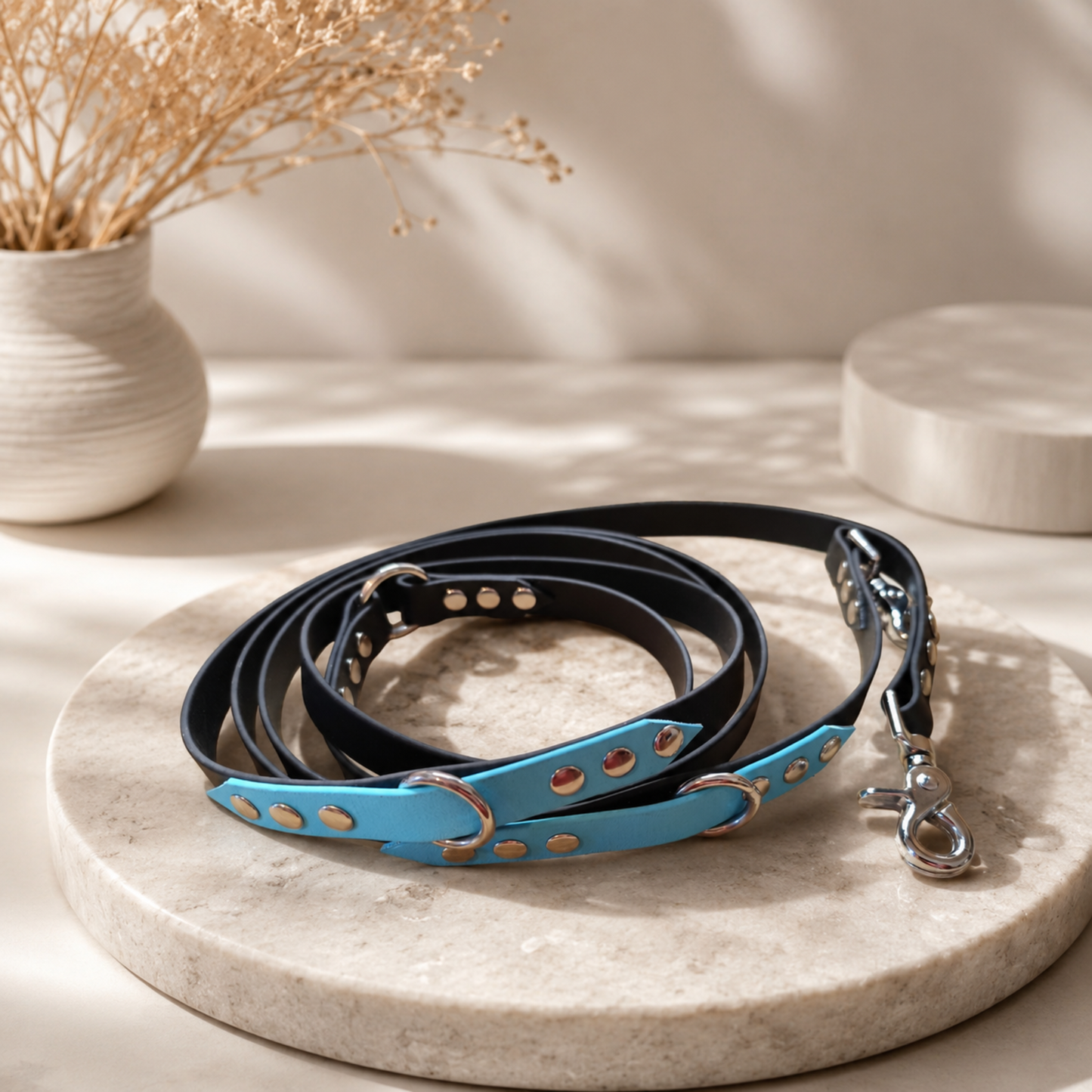 Laisse multiposition en BioThane noir et bleu ciel avec bouclerie inox, présentée sur un support en pierre dans un décor minimaliste.
